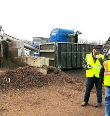 Two people stand in front of a large machine at a compost processing facility in Maryland.